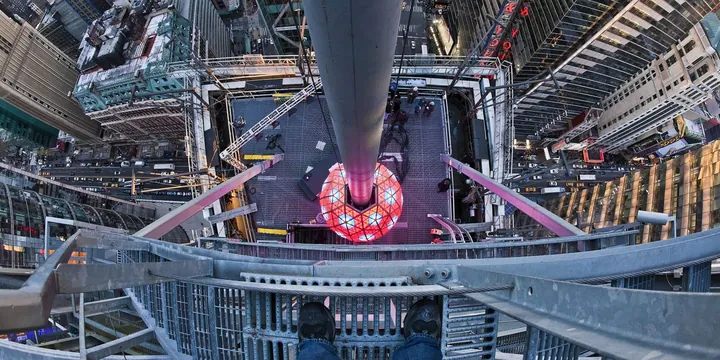 Looking straight down from the top of the Times Square flagpole at the New Year's Eve ball, with a person's sneakers on the steel platform and the crowded streets far below