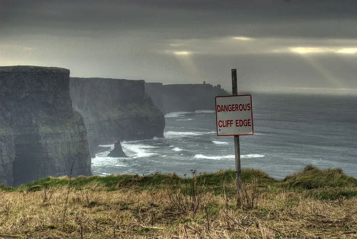 Red-and-white "Dangerous Cliff Edge" sign on a grassy clifftop above dark sea cliffs and crashing waves under a stormy sky.